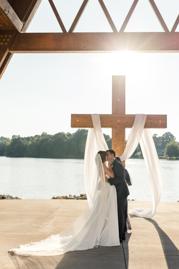 Bride in white gown and veil kissing groom in blue suit in front of a giant wooden cross photographed by Charlotte Wedding Photographer