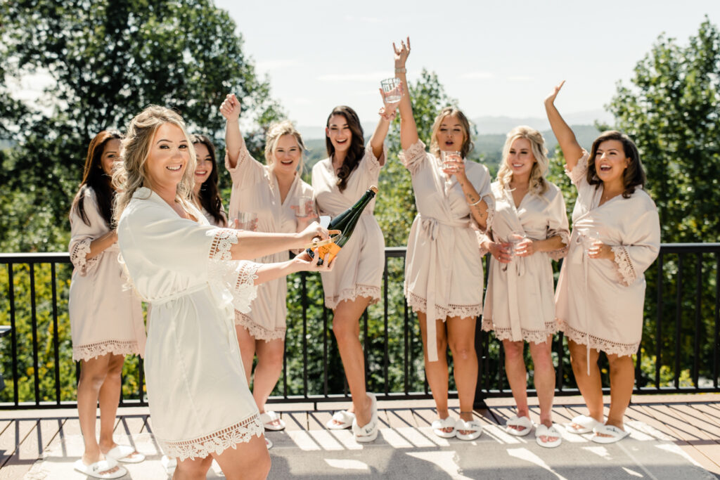 Bride with long blonde hair in a white robe popping champagne bottle with seven of her bridesmaids wearing pink robes in Asheville. Photographed by Charlotte wedding photographer.
