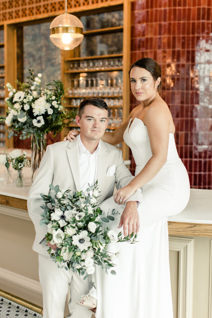 Bride, in white strapless dress, sitting on bar counter with arm around her groom, in a tan suit with white and green flowers at The Ruth wedding venue in Charlotte NC. Photographed by Charlotte Wedding Photographer.