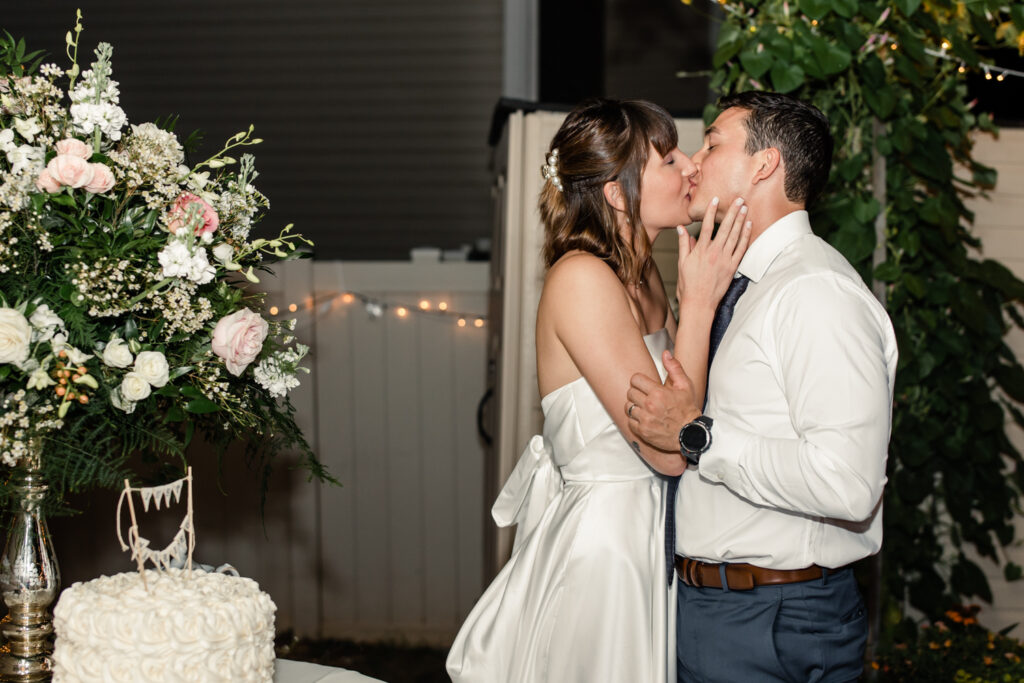 Bride in white gown and kissing groom in white button down and blue tie next to white wedding cake photographed by Charlotte Wedding Photographer