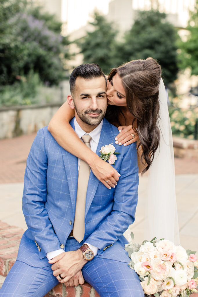 Bride in white dress with brown hair hugging and kissing cheek of the groom in blue suit in uptown Charlotte photographed by Charlotte Wedding Photographer.  