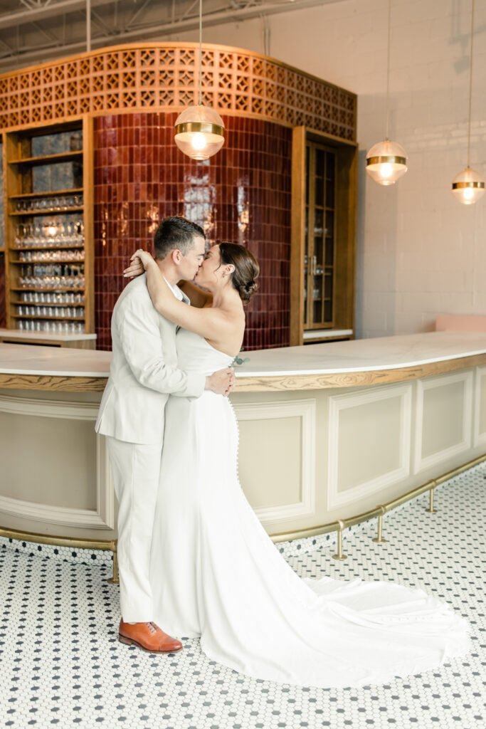Bride, in white strapless dress, kissing her groom, in tan suit, in front of bar at The Ruth wedding venue in Charlotte NC. Photographed by Charlotte Wedding Photographer.