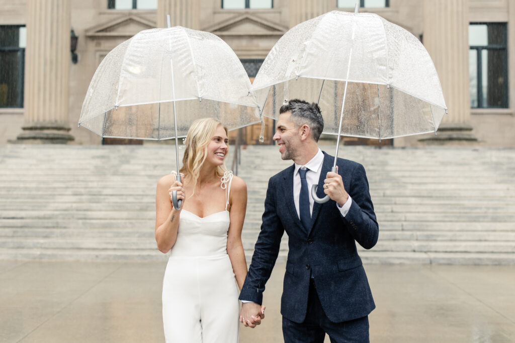 Bride, with blonde hair in white jumpsuit, and groom, white grey hair in navy suit, walking under a clear umbrella. Photographed by Charlotte Wedding Photographer.