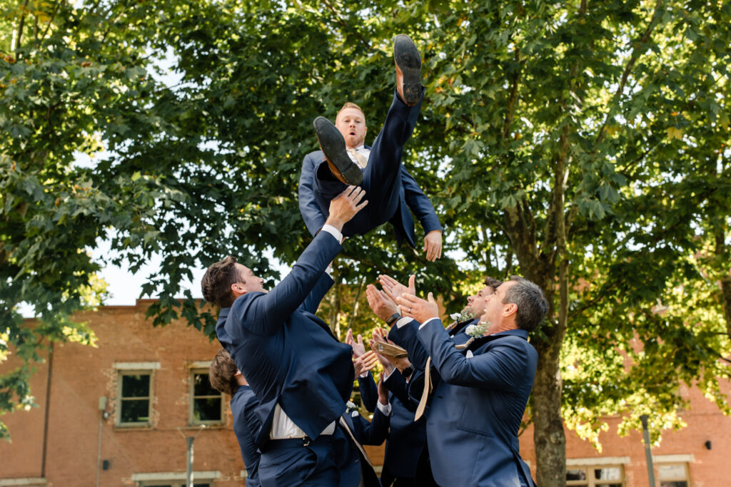 Groomsmen in blue suits throwing the groom in a blue suit in the air. Photographed by Charlotte wedding photographer.