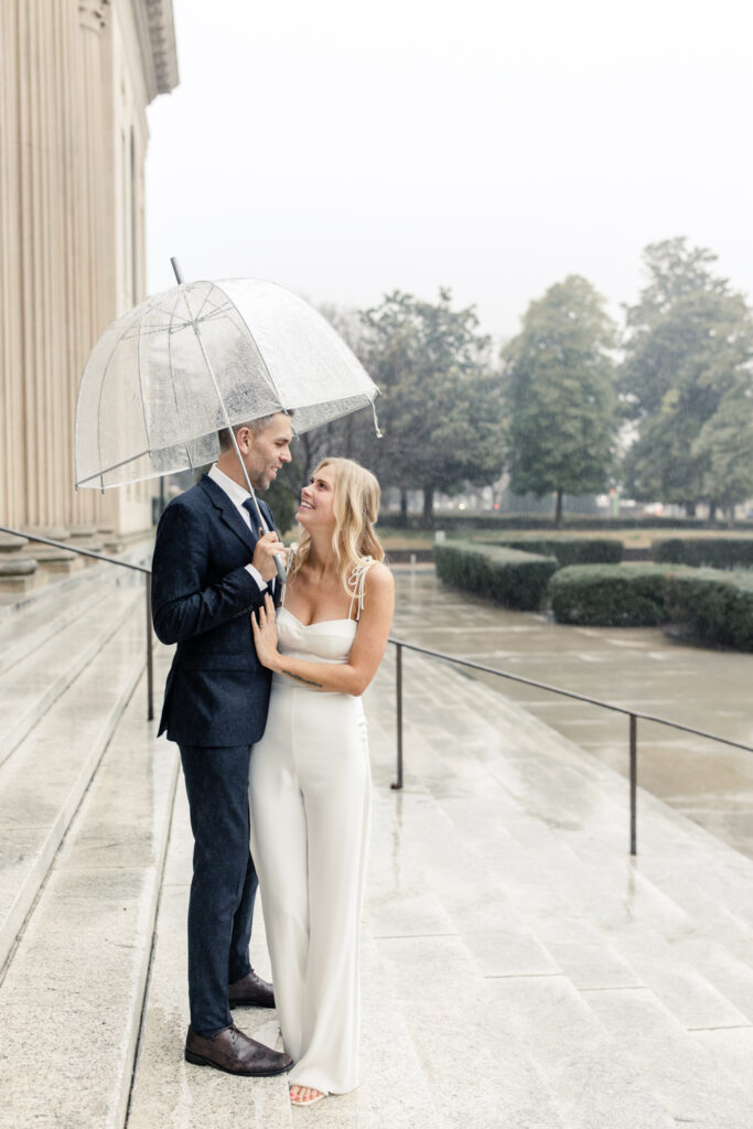 Groom, with grey hair in navy suit, smiling at his bride, in white jumpsuit,  under a clear umbrella in uptown Charlotte NC. Photographed by Charlotte Wedding Photographer.