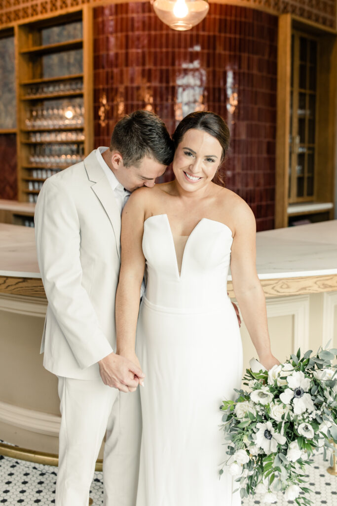 Groom, in tan suit, kissing the shoulder of his bride, in white strapless dress at The Ruth wedding venue in Charlotte NC. Photographed by Charlotte Wedding Photographer.