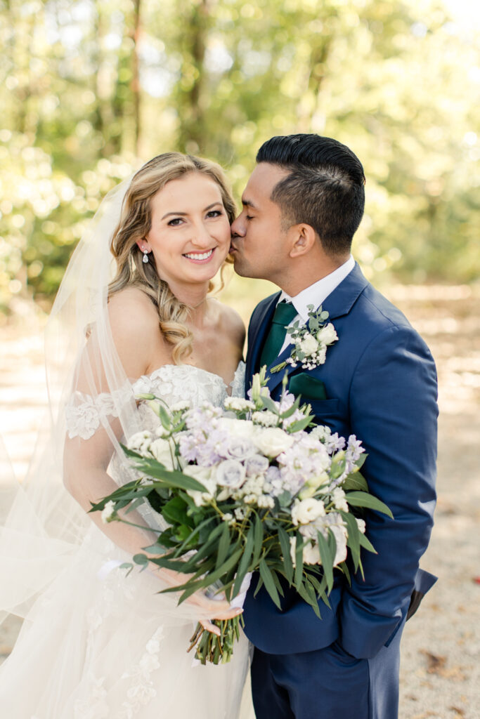 Groom in blue suit kissing the cheek of bride in white lace gown holding purple and white floral bouquet at Riverwood Manor Wedding Venue. Photographed by Charlotte wedding photographer, Stephanie Bailey.