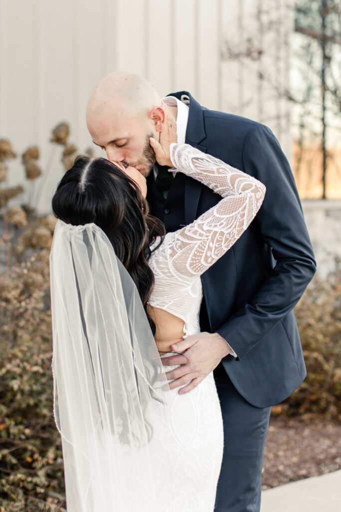 Bride in white long sleeve less dress and veil kissing her groom in blue suit at Fields at Skycrest Wedding Venue in Charlotte NC. Photographed by Charlotte wedding photographer.
