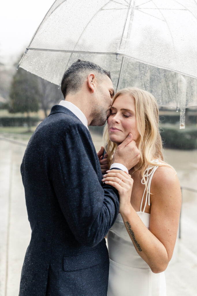 Groom, with grey hair in navy suit, kissing the cheek of blonde bride under a clear umbrella in uptown Charlotte NC. Photographed by Charlotte Wedding Photographer.