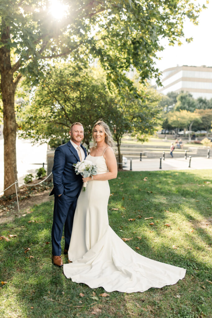 Bride in white lace dress with long blonde hair standing with groom in blue suit with a gold tie in Asheville NC with green trees in the background. Photographed by Charlotte wedding photographer.