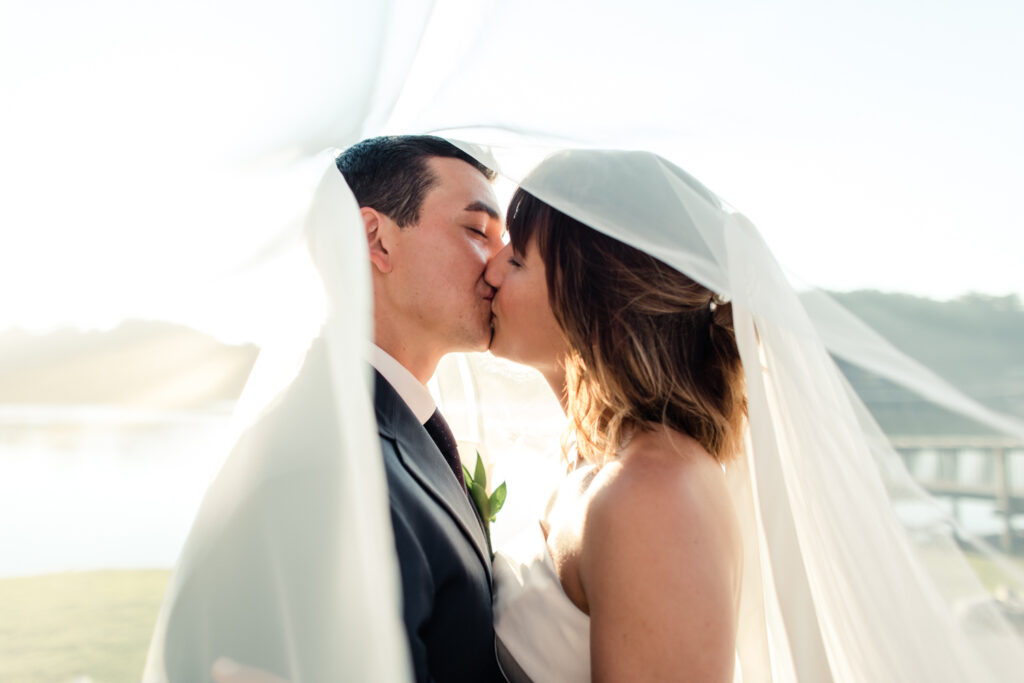 Bride in white gown and veil kissing groom in blue suit under white veil photographed by Charlotte Wedding Photographer