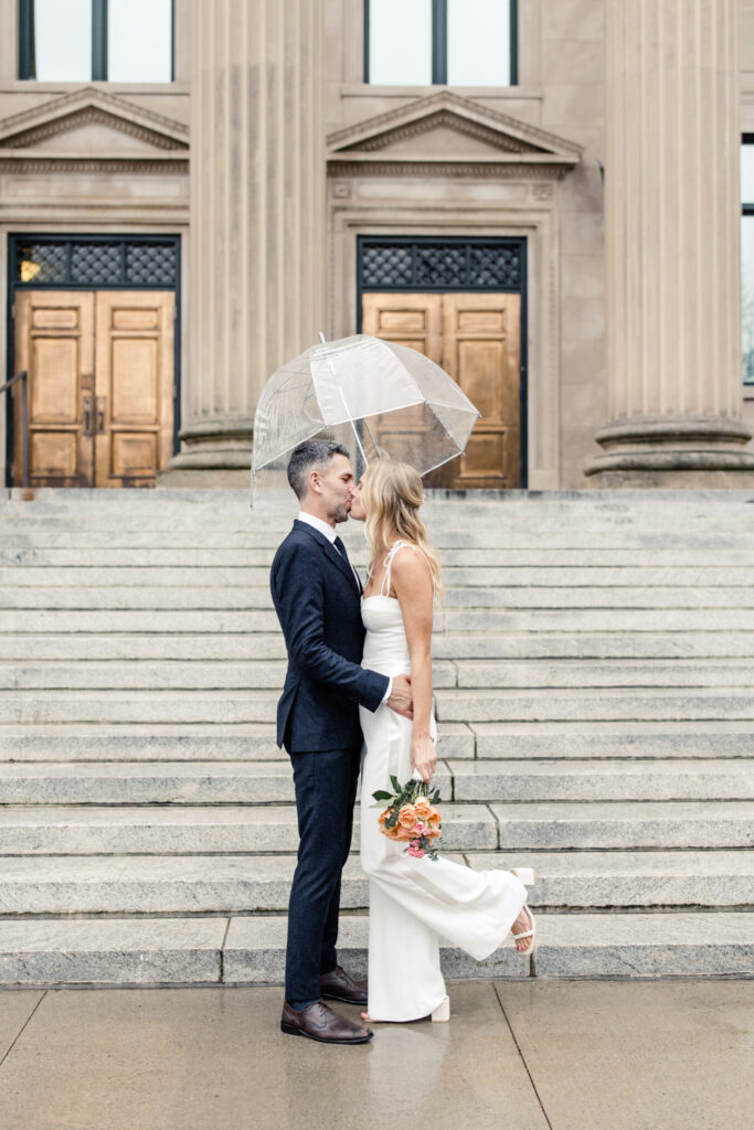 Bride, with blonde hair in white jumpsuit, and groom, white grey hair in navy suit, kissing under a clear umbrella. Photographed by Charlotte Wedding Photographer.