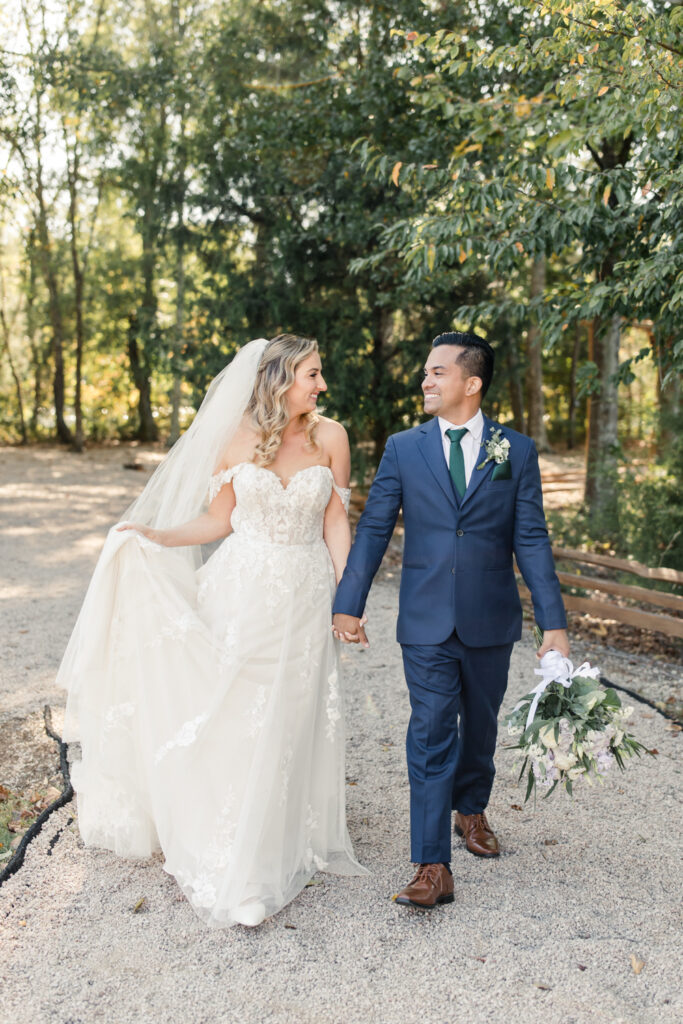 Bride with long blonde hair in a white lace dress and long veil walking holding hands with groom in blue suit at Riverwood Manor Wedding Venue. Photographed by Charlotte wedding photographer, Stephanie Bailey.