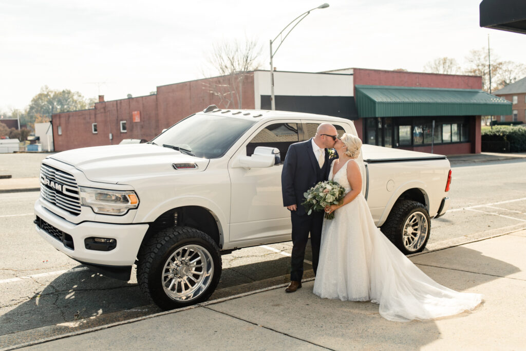 Bride with blonde hair in a white lace dress kissing her groom, in a navy suit and sunglasses in front of a white pick up truck at The Bottle Factory Wedding Venue. Photographed by Charlotte wedding photographer. 