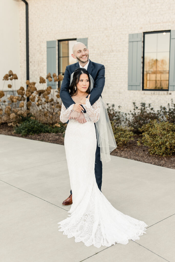 Bride in white long sleeve less dress and veil smiling and hugging with her groom in blue suit at Fields at Skycrest Wedding Venue in Charlotte NC. Photographed by Charlotte wedding photographer.