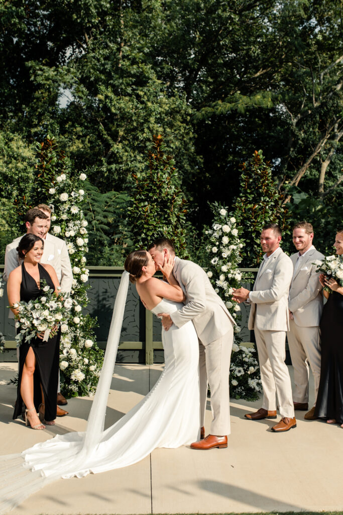 Bride, in white strapless dress, kissing her groom, in tan suit, in front of white and green floral arbor at The Ruth wedding venue in Charlotte NC. Photographed by Charlotte Wedding Photographer.
