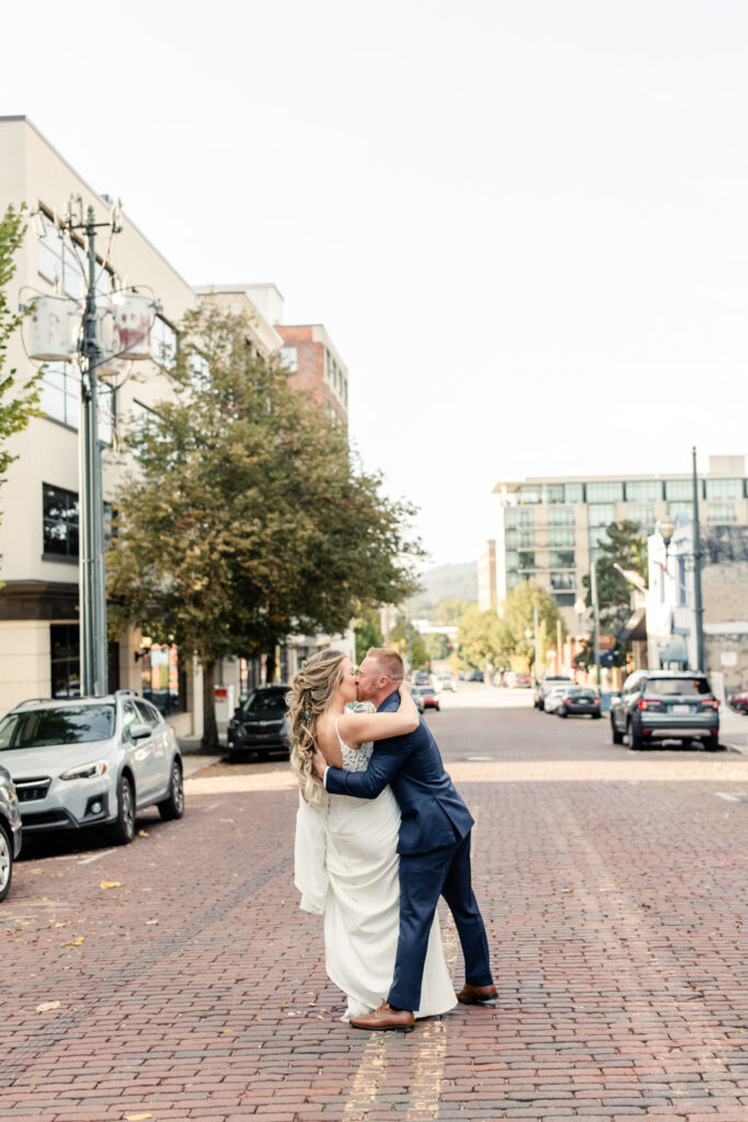 Bride in white dress with long blonde hair kissing her groom in a blue suit in the middle of the street on cobble stone at The Venue in Asheville NC. Photographed by Charlotte wedding photographer.
