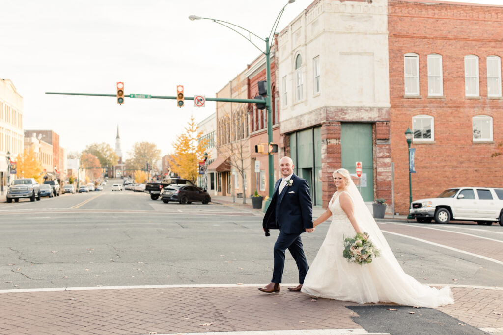 Bride with blonde hair in a white lace dress holding hands with groom, in a navy suit in the street at The Bottle Factory Wedding Venue. Photographed by Charlotte wedding photographer. 