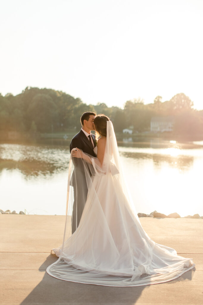 Bride in white gown and veil kissing groom in blue suit in front lake photographed by Charlotte Wedding Photographer