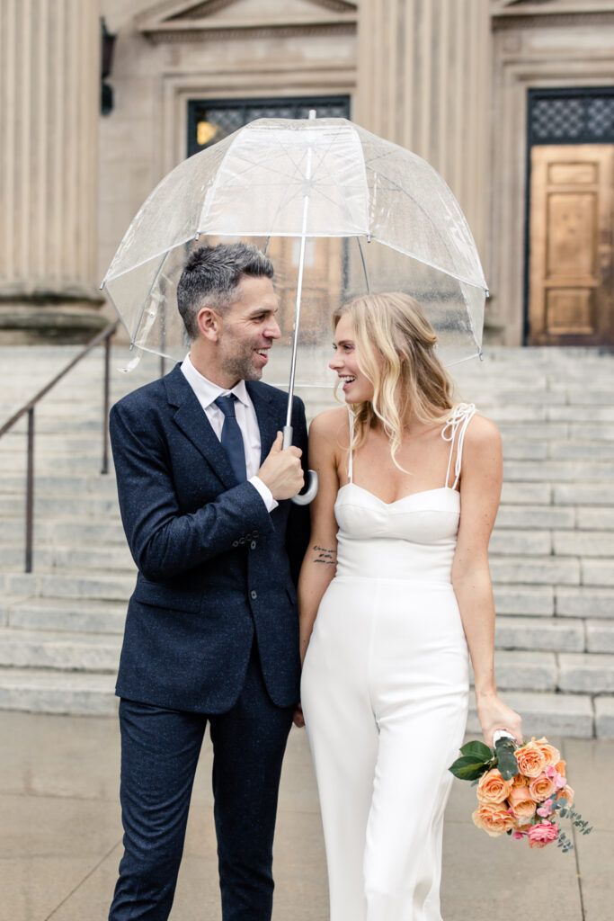 Bride, with blonde hair in white jumpsuit, and groom, white grey hair in navy suit, walking under a clear umbrella. Photographed by Charlotte Wedding Photographer.