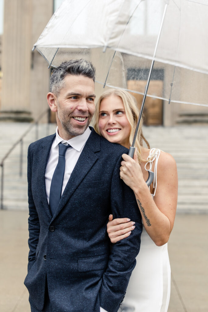 Bride, with blonde hair in white jumpsuit, hugging her groom, white grey hair in navy suit, under a clear umbrella. Photographed by Charlotte Wedding Photographer.