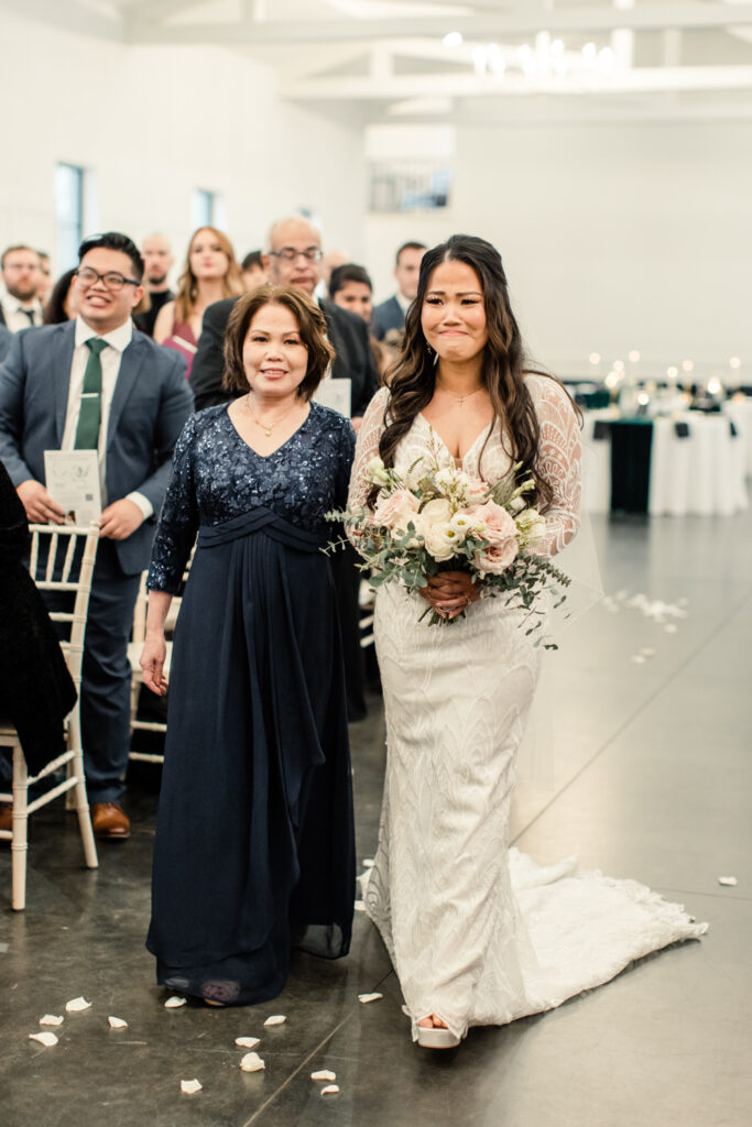 Bride in white long sleeve lace dress walking down ceremony aisle with her mom in a navy dress at Fields at Skycrest Wedding Venue in Charlotte NC. Photographed by Charlotte Wedding photographer.