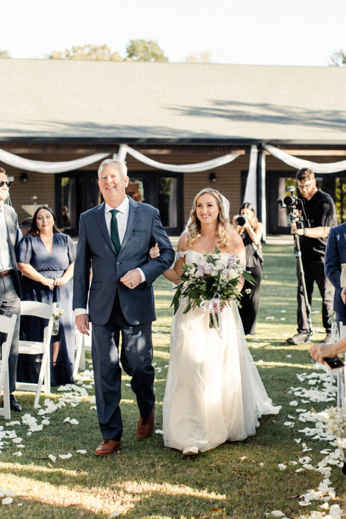 Bride in white lace wedding dress holding white, purple and green floral bouquet walking walking down ceremony aisle with father in blue suit at Riverwood Manor Wedding Venue. Photographed by Charlotte wedding photographer, Stephanie Bailey. 