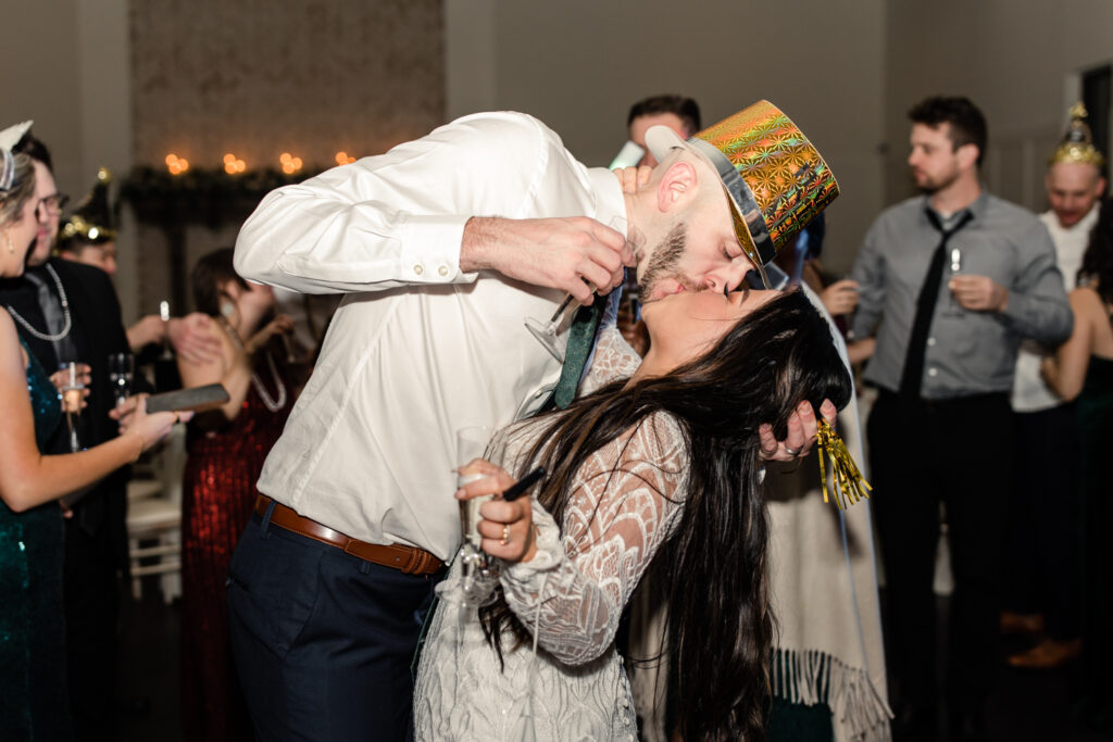 Bride with dark brown hair in a long white lace dress kissing groom in white button down with green tie on new years eve during reception at Fields at Skycrest Wedding Venue in Charlotte NC. Photographed by Charlotte wedding photographer.
