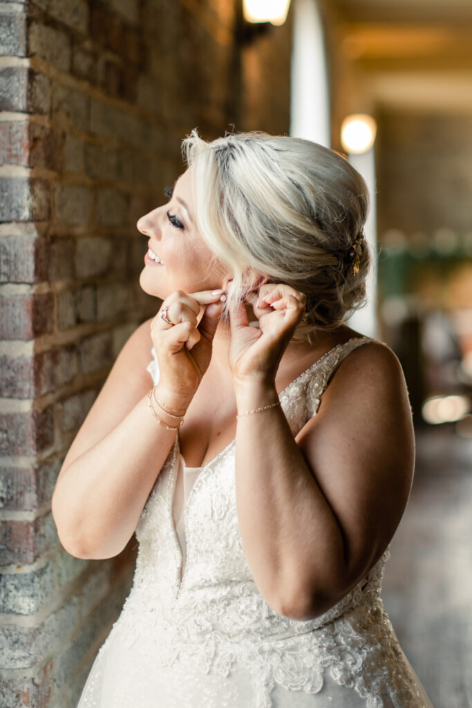 Bride with blonde hair in a white lace dress putting on her earrings at The Bottle Factory Wedding Venue. Photographed by Charlotte wedding photographer. 