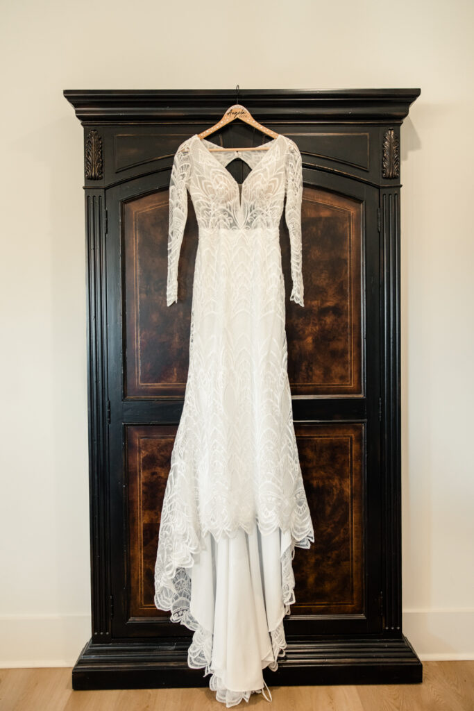 White lace long sleeve wedding dress hanging on a dark brown wood wardrobe closet at Fields at Skycrest Wedding Venue in Charlotte NC. Photographed by Charlotte wedding photographer.